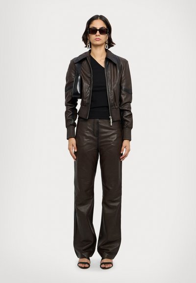 Blouson en cuir marron avec une fermeture éclair à l'avant, des poignets côte à côte et un pantalon large en cuir marron assorti, associé à des sandales noires.