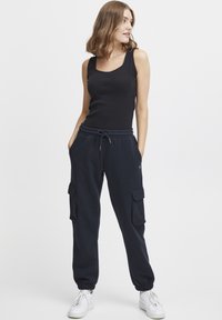 Zwarte geribbelde tanktop gecombineerd met marineblauwe trainingsbroek met cargozakken, elastische boorden en een trekkoord in de taille. Model draagt witte sneakers.