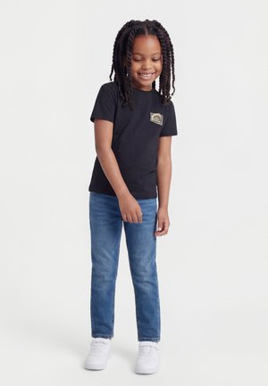 Noor laps palmikutega, kes kannab musta t-särki, siniseid teksapükse ja valgeid tossusid, seisab ning naeratab ühevärvilise tausta ees.