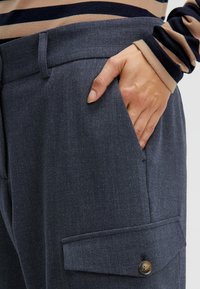 Dunkelgraue maßgeschneiderte Hose mit strukturierter Oberfläche, ausgestattet mit Seitentaschen und einer Knopfverschluss-Tasche für zusätzliche Details.