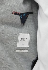 Világosszürke, textúrált anyagú dzseki belső. Címkék: "NEXT TAILORING", 40R méret, és "SLIM FIT." Virágmotívum a zsebben.