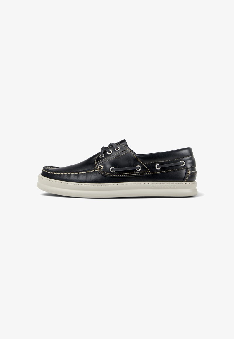 Black leather boat shoe with a white rubber sole, featuring decorative stitching and metal eyelets for laces. Classic design with minimal accents.