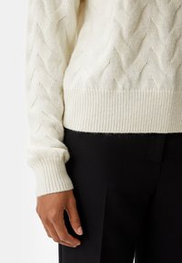 Un pull en tricot couleur crème avec un motif en câble, des poignets et un ourlet côtelés, associé à un pantalon noir ajusté.