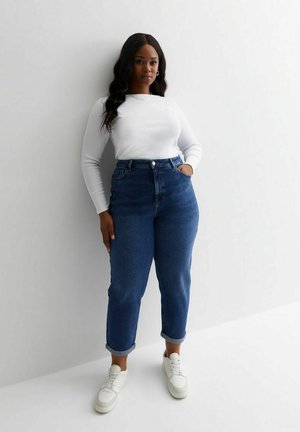 Jeans in denim blu a vita alta con una vestibilità rilassata, con risvolti, abbinati a un top a maniche lunghe bianco attillato e sneakers bianche.