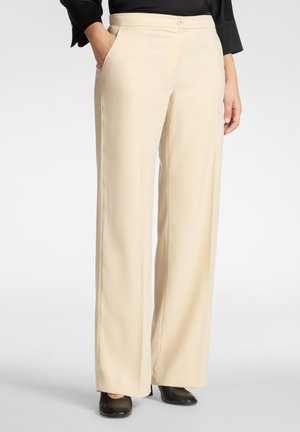 Frau trägt beige weite Hosen mit Seitentaschen und schwarzen Absatzschuhen, steht mit einer Hand in der Tasche vor einfarbigem Hintergrund.