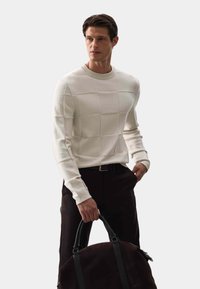 Cremfarbener Pullover mit Karomuster, aus strukturiertem Stoff. Getragen mit dunklen Hosen und hält eine braune Tasche mit schwarzen Akzenten.