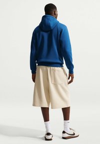 Mann steht mit dem Rücken zur Kamera, trägt einen blauen Kapuzenpullover, beige knielange Shorts mit einer Gesäßtasche, weiße Socken und weiße Nike-Sneaker.