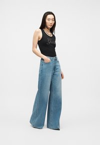 Femme debout, une main dans la poche, portant un haut noir sans manches de la marque Guess et un jean large bleu, sur fond blanc.