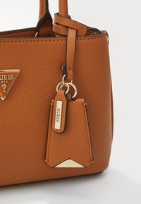 Borsa a tracolla in pelle marrone con texture, logo metallico triangolare, etichetta decorativa e accenti in metallo dorato. Design strutturato, cuciture senza soluzione di continuità.