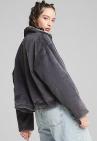 Giacca grigia in peluche, corta, con colletto alto, vestibilità ampia e maniche lunghe. Il tessuto strutturato appare morbido e caldo.