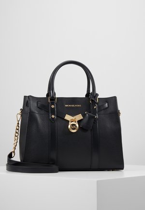 Schwarze Michael Kors Handtasche mit zwei Griffen, goldenem Logohänger, Kettendetail am Riemen und genarbter Lederstruktur auf weißer Oberfläche.