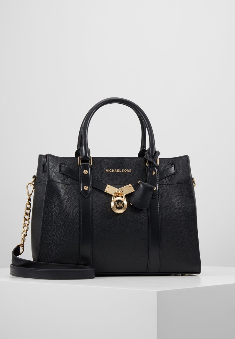 Sac à main Michael Kors noir avec doubles poignées, breloque logo dorée, détail chaîne sur la sangle et texture cuir grainé sur une surface blanche.