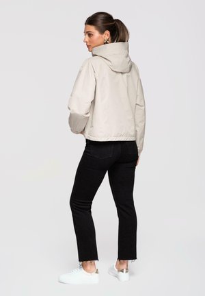 Femme avec queue de cheval portant une veste à capuche beige, un jean court noir et des baskets blanches, debout de profil sur un fond uni.