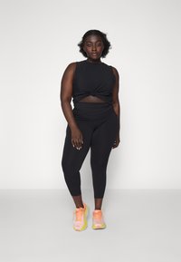Top corto atlético negro con detalle anudado, combinado con leggings ajustados negros. La modelo lleva zapatillas deportivas multicolores con acentos naranjas.