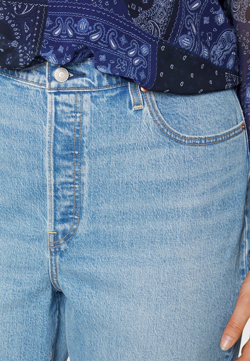 Hellblaue Jeans mit hoher Taille, featuring Kupferbeschläge, sichtbare Nahtdetails und Texturvariationen. Ein gemusterter dunkler Oberteil ist teilweise sichtbar.