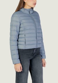 Giacca a piumino azzurro chiaro con design trapuntato orizzontale, chiusura con zip e collo alto. Indossata con jeans grigio scuro, le maniche sono a lunghezza intera.