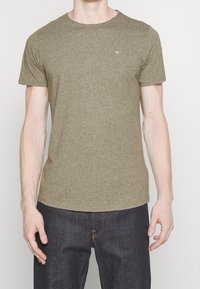 Muž v jednobarevném olivově zeleném tričku s krátkým rukávem a tmavě modrých džínách, stojící na jednobarevném světlém pozadí.