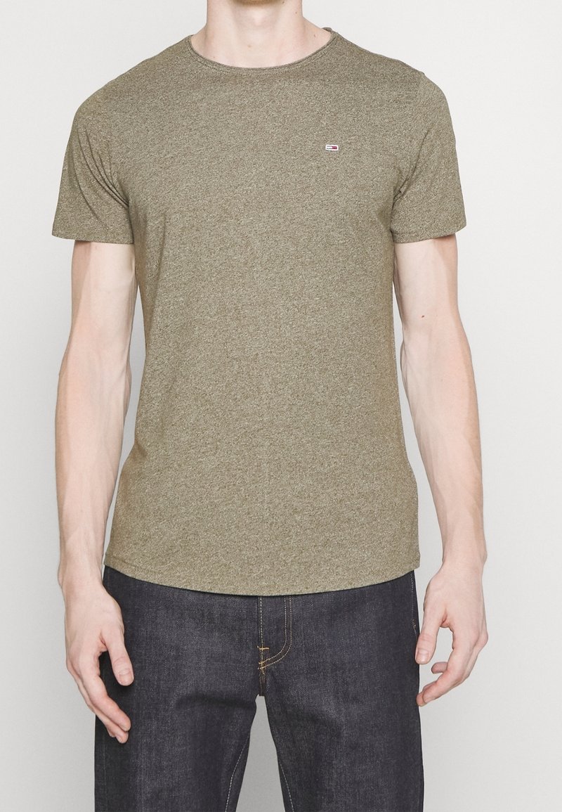 Muž v jednobarevném olivově zeleném tričku s krátkým rukávem a tmavě modrých džínách, stojící na jednobarevném světlém pozadí.