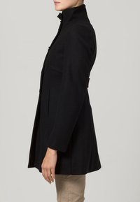 Svart ullkappa med hög krage, skräddarsydd passform, sidofickor och en slät yta. Den figursydda designen framhäver silhuetten utan utsmyckningar.