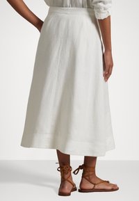 Falda midi de lino blanco con silueta acampanada, que presenta una cintura lisa y una abertura lateral. Combinada con sandalias marrones de tiras.