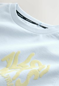 Světle modré bavlněné tričko s výrazným žlutým grafickým textovým motivem. Výstřih je v klasickém crew stylu s žebrovanou texturou.