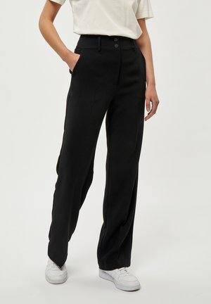Femme portant un pantalon noir taille haute à jambes larges avec boutons sur le devant, poches latérales, baskets blanches et un top blanc à manches courtes rentré.