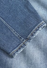 Középkék farmeranyag, amely szakadt mandzsettával, látható aranysárga varrásokkal és sima textúrával rendelkezik, enyhe kopással.