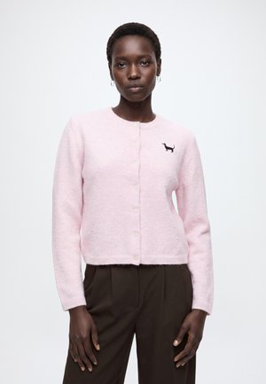 Femme portant un cardigan rose clair boutonné avec une petite broderie noire de chien sur la poitrine et un pantalon marron foncé.