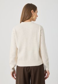 Žena s dlouhými hnědými vlasy, oblečená v krémovém svetru se perlovými a korálkovými detaily na rukávech a v tmavě hnědých kalhotách, otočená zády.