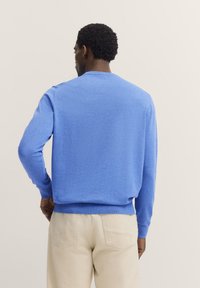 Blauer Rundhalsausschnitt-Pullover mit langen Ärmeln, weichem Stoff, gerippten Bündchen und Saum, minimalistischem Design, über beigen Hosen getragen, von hinten betrachtet.