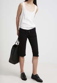 Kobieta w białej bluzce bez rękawów, czarnych spodniaczkach, czarnych butach wsuwanych, trzymająca dużą czarną torbę na tle jednolitym.