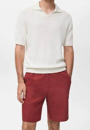 Homme portant un polo en maille blanche texturée à manches courtes et un short rouge ajusté jusqu’à mi-cuisse, les mains dans les poches.