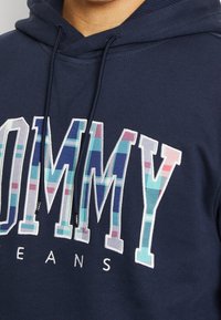Sudadera azul marino con un gran logo "TOMMY" en un patrón de cuadros multicolor, con bordado blanco y cordones negros. La tela parece suave.