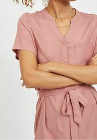 Krátký rukáv, zapínací šaty z měkké růžové tkaniny. Obsahuje límeček, náprsní kapsu a zavázaný pas pro přiléhavou siluetu.