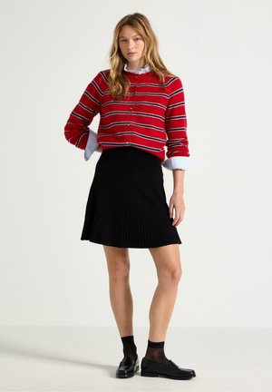 Kvinde iført en rødstribet sweater, sort plisseret nederdel, sorte sokker og sorte loafers, stående mod en ensfarvet hvid baggrund.