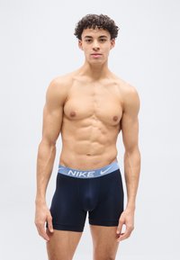 Νέος άνδρας με σγουρά μαλλιά που φοράει μαύρα ανδρικά εσώρουχα Nike με ανοιχτό μπλε ζώνη μέσης, πάνω σε απλό λευκό φόντο.
