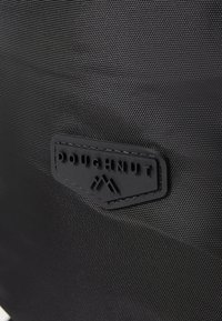 Sort stofpose med en tekstureret overflade. En sort gummimærke med prægede bogstaver og logo er tydeligt præsenteret.