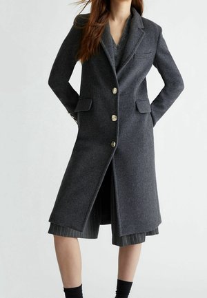 Kvinde iført en mørkegrå uldfrakke til knæet med tre knapper, spids krave, klaplommer, over en grå stribet nederdel og sorte sokker.