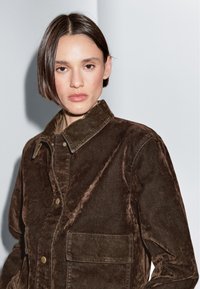 Jonge vrouw met kort donker haar draagt een donkerbruine ribfluwelen jas met grote voorzak, staand tegen een lichtgrijze achtergrond.