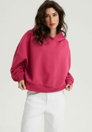 Kobieta o ciemnych falowanych włosach ubrana w różową, za dużą bluzę z kapturem i białe spodnie, trzymająca brzeg bluzy, stojąca na jednolitym jasnym tle.