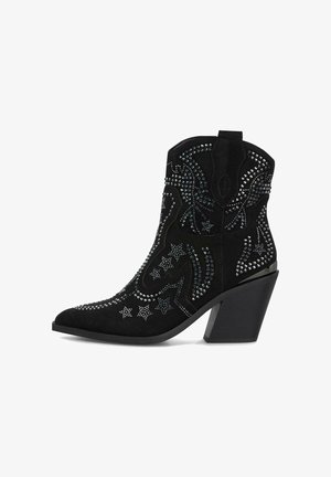 Schwarze Wildleder-Ankelboots mit spitzem Zehenbereich, verziert mit metallischen Nieten, Sternmustern und einem massiven Absatz. Detaillierte Nahtarbeit am Schaft.