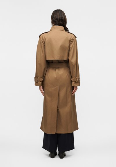 Γυναίκα που φοράει ένα μακρύ καφέ παλτό τύπου τσέστερφιλντ με ζώνη και λεπτομέρειες με αγκράφα, στέκεται με την πλάτη γυρισμένη, με μαύρο παντελόνι και ορατά παπούτσια με τακούνι.