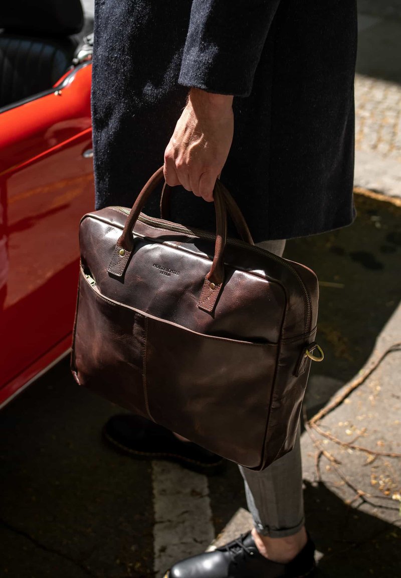 Man in dark coat holding brown leather briefcase near red car, standing on street with sunlight and shadows.