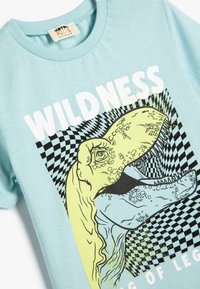 Lyseblå bomulds-t-shirt med et grafisk design af et dinosaurus-hoved i grøn og sort, med en ternet baggrund og fed tekst.
