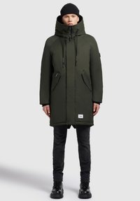 Man in een donkergroene hooded winterjas, zwarte beanie, zwarte jeans en zwarte laarzen, staand tegen een effen grijze achtergrond.