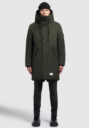 Mand iført mørkegrøn vinterfrakke med hætte, sort hue, sorte jeans og sorte støvler, der står foran en ensfarvet grå baggrund.