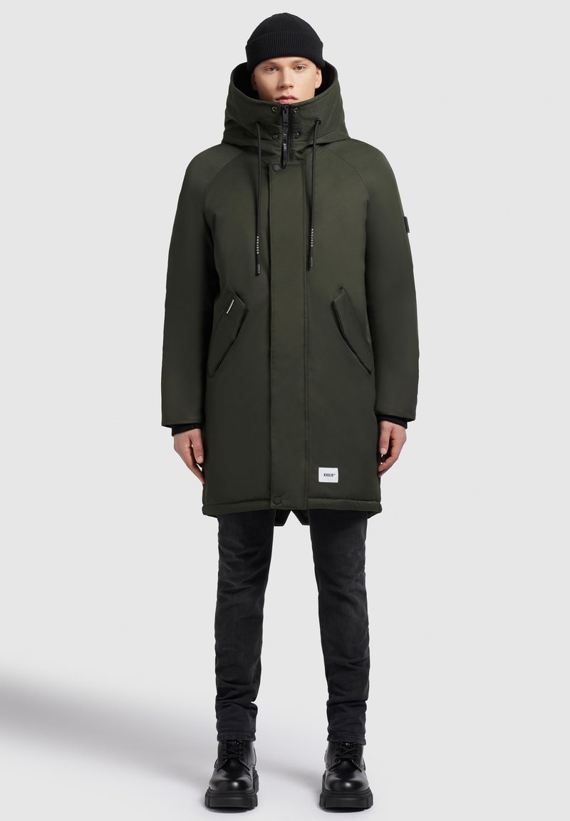 Man in een donkergroene hooded winterjas, zwarte beanie, zwarte jeans en zwarte laarzen, staand tegen een effen grijze achtergrond.