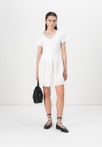 White short dress with a ribbed bodice and embroidered skirt, featuring a scalloped hem. Model holds a black bag and wears black lace-up flats.