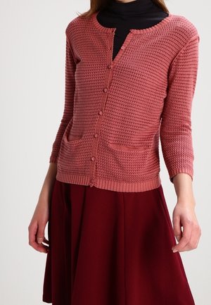 Cardigan rosa chiaro con maniche a tre quarti, maglia testurizzata, scollo rotondo, tasche frontali e bottoni al centro, abbinato a una gonna bordeaux.