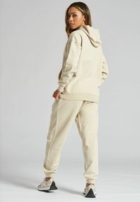 Beżowa bluza z kapturem i dresy wykonane z miękkiego materiału, o luźnym kroju, z ribbowanymi ściągaczami i bocznymi kieszeniami. Obuwie ma jasny kolor i teksturowaną podeszwę.